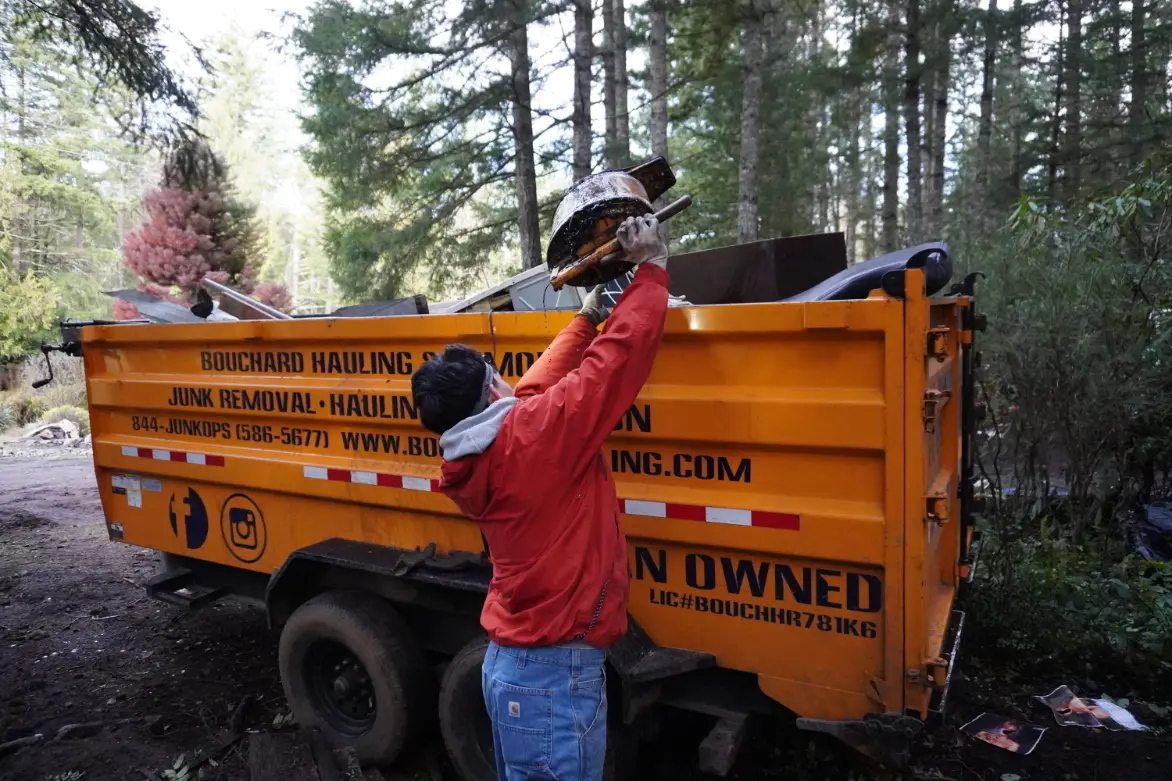 A person in a red jacket loading a dirty helmet or object into the back of an orange dumpster truck labeled &ldquo;BOUCHARD HAULING&rdquo; in a wooded area.